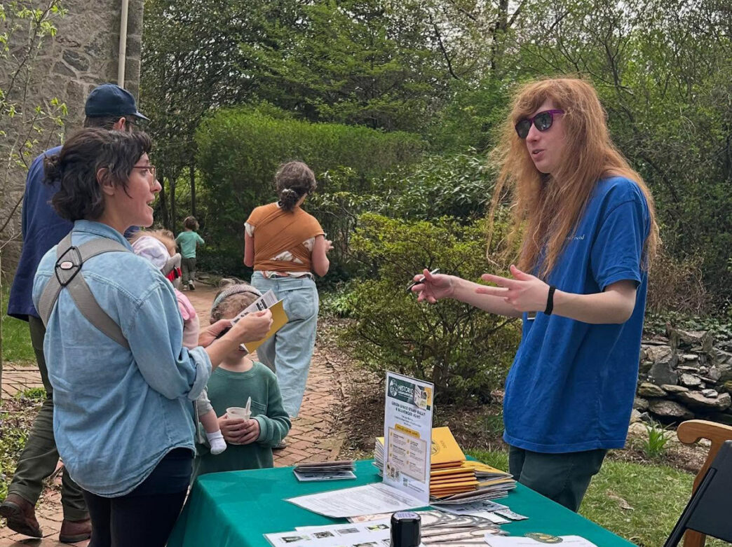 A Germantown Green Space Stamp Rally participant discusses the event with an organizer outdoors with lush greenery and other participants in the background.