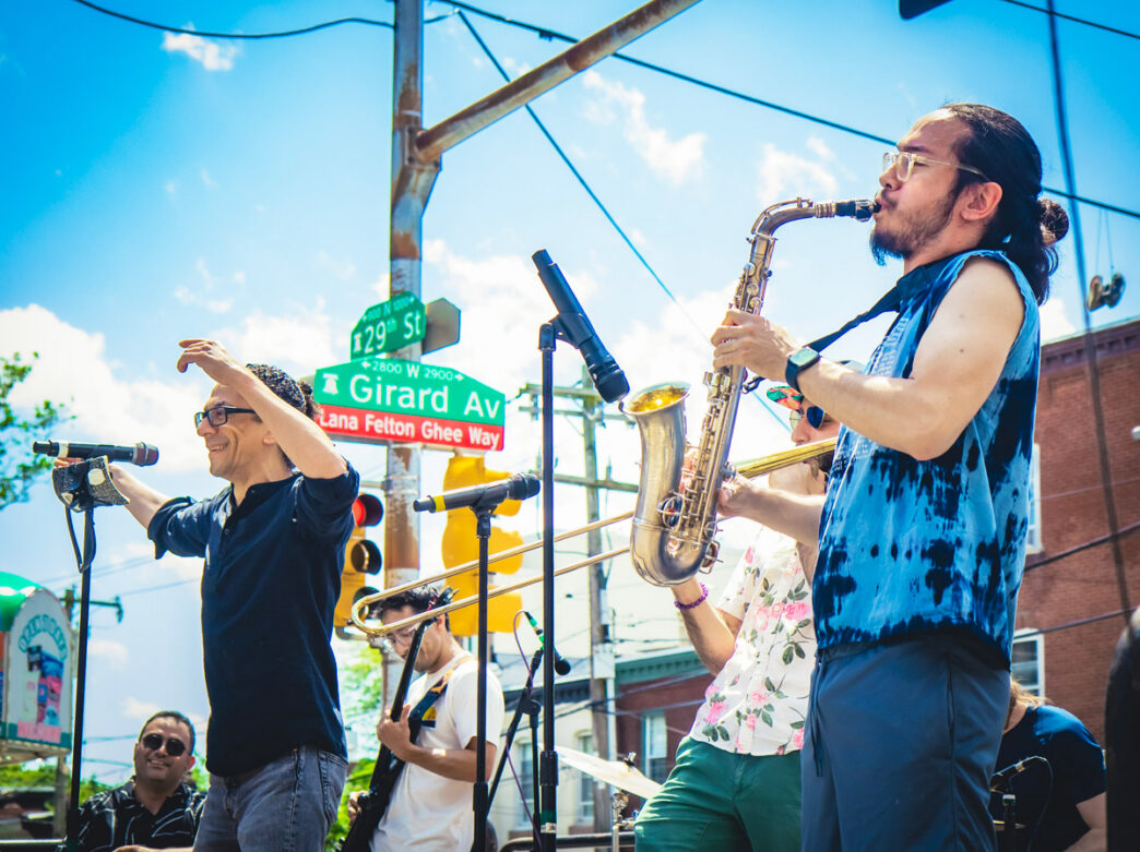 A saxophonist and singer perform with a band at the Girard Avenue Street Fest in Brewerytown, with the street signs for 29th Street and Girard Avenue in the background.
