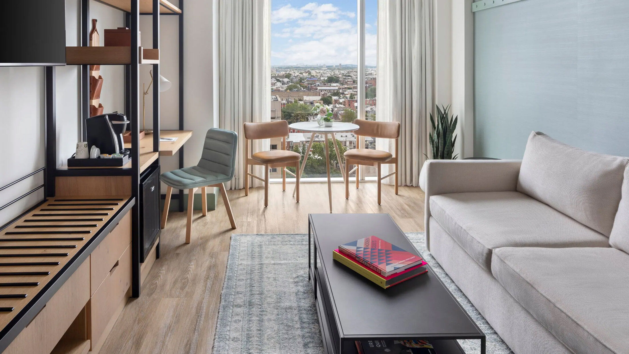 Modern guest room at Hyatt Centric Philadelphia featuring a light-filled seating area with a sofa, coffee table, and small dining set by a large window overlooking the city skyline.