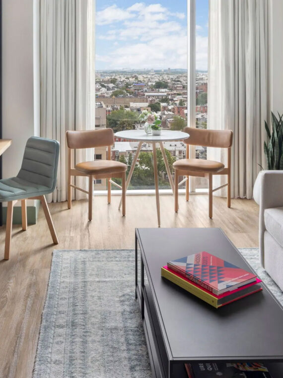 Modern guest room at Hyatt Centric Philadelphia featuring a light-filled seating area with a sofa, coffee table, and small dining set by a large window overlooking the city skyline.