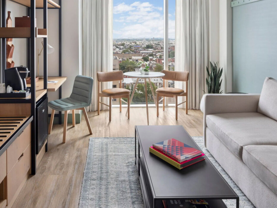 Chambre moderne du Hyatt Centric Philadelphia comprenant un coin salon lumineux avec un canapé, une table basse et une petite table à manger près d'une grande fenêtre donnant sur la ligne d'horizon de la ville.