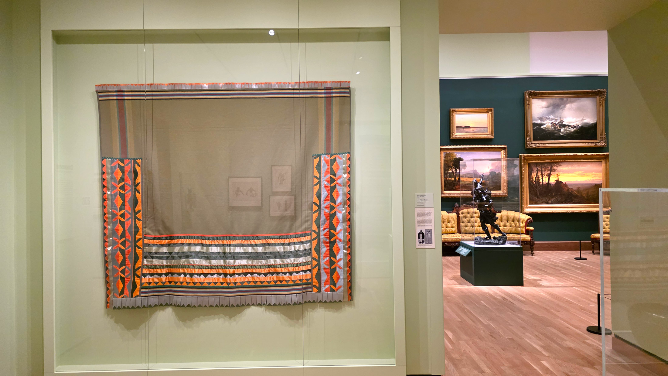 Textile with geometric patterns in orange, gray, and black displayed behind glass in Philadelphia’s A Nation of Artists exhibit, with a view into an adjacent gallery featuring framed landscape paintings and a sculpture.