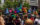 Dancers performing at Philadelphia's Juneteenth Block Party on an outdoor stage, wearing vibrant colorful costumes in shades of blue, red, yellow, and orange, with a crowd of spectators watching below and festive floral decorations and greenery surrounding the stage.