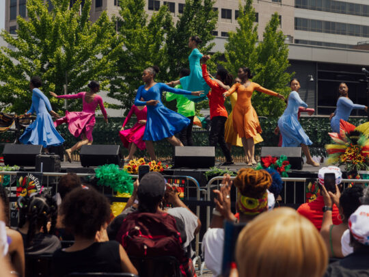 Bailarines actuando en la fiesta callejera del Juneteenth de Filadelfia, en un escenario al aire libre, con trajes de vivos colores en tonos azules, rojos, amarillos y naranjas, ante una multitud de espectadores que los observan desde abajo y rodeados de festivas decoraciones florales y vegetación.