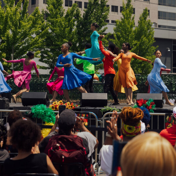 Des danseurs se produisent lors de la Juneteenth Block Party de Philadelphie sur une scène en plein air, vêtus de costumes aux couleurs vives dans les tons bleu, rouge, jaune et orange, sous le regard d'une foule de spectateurs. Des décorations florales festives et de la verdure entourent la scène.