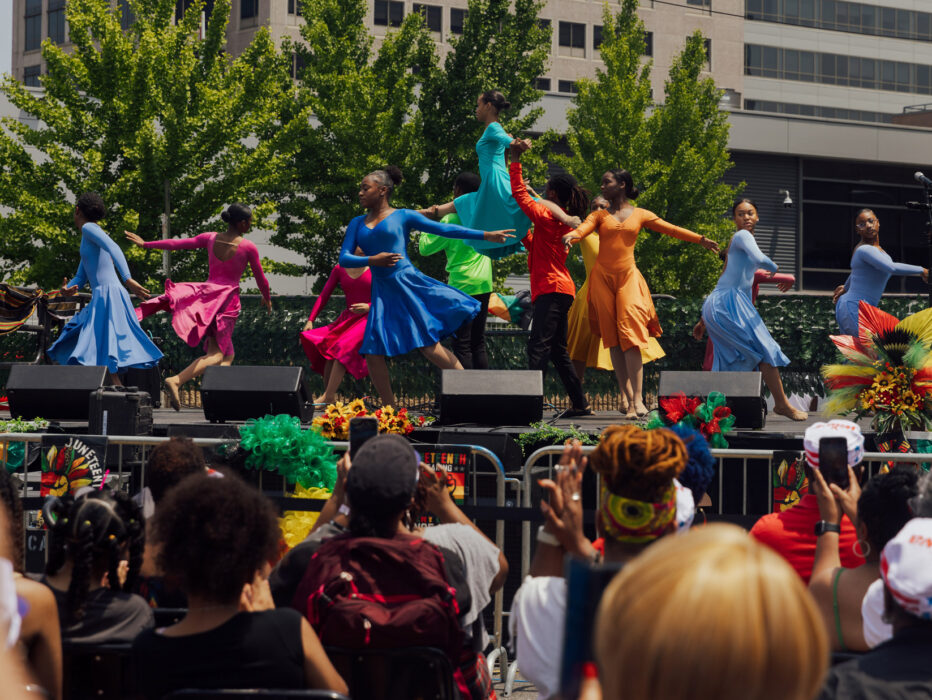 Dancers performing at Philadelphia's Juneteenth Block Party on an outdoor stage, wearing vibrant colorful costumes in shades of blue, red, yellow, and orange, with a crowd of spectators watching below and festive floral decorations and greenery surrounding the stage.