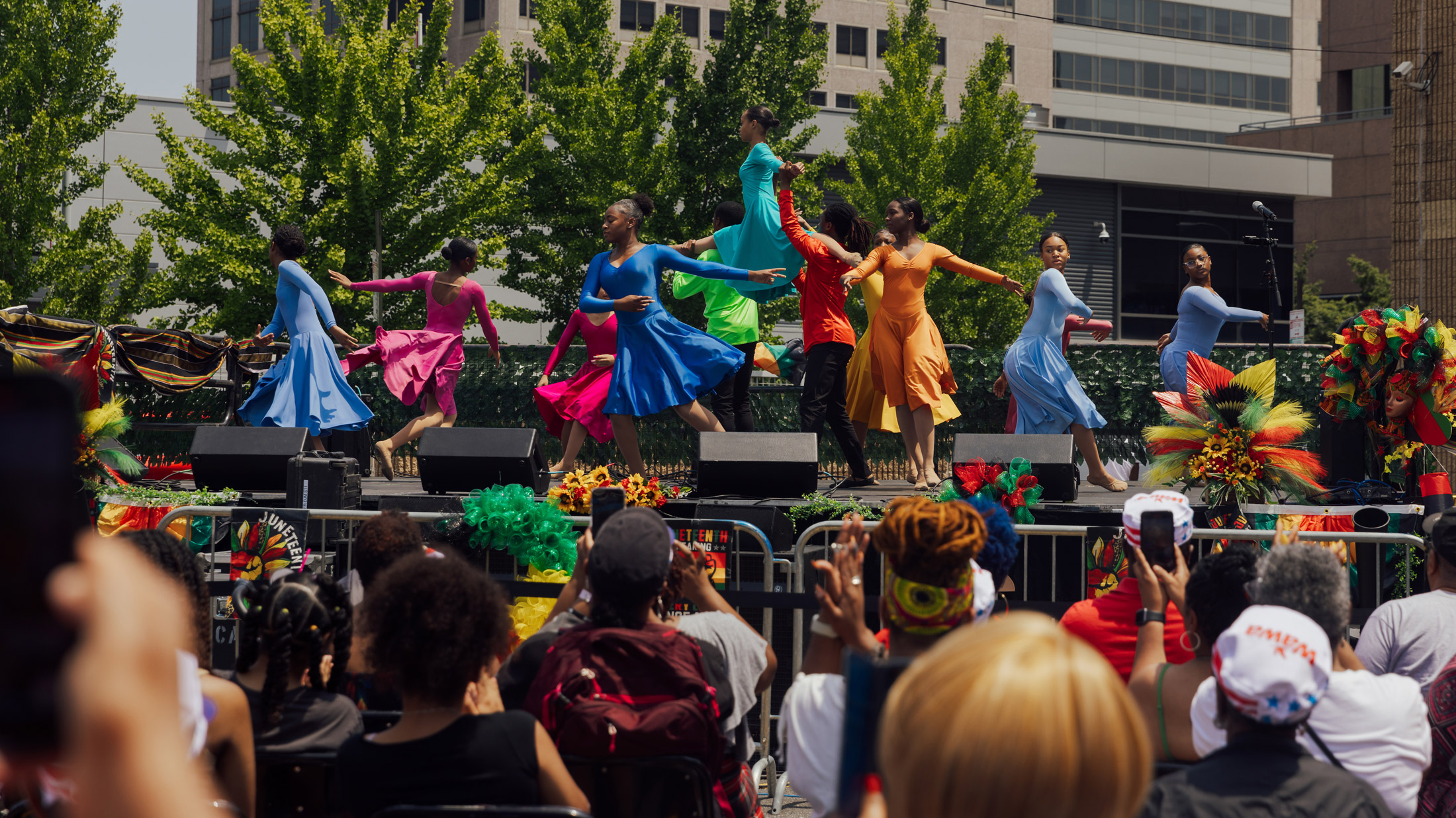 Dancers performing at Philadelphia's Juneteenth Block Party on an outdoor stage, wearing vibrant colorful costumes in shades of blue, red, yellow, and orange, with a crowd of spectators watching below and festive floral decorations and greenery surrounding the stage.