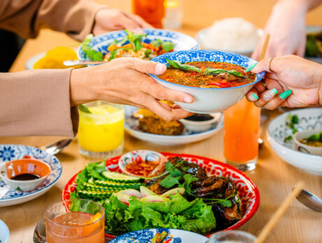 Diners at Kalaya in Philadelphia pass a bowl of vibrant Thai curry across a table filled with colorful dishes, fresh herbs, and bright cocktails.