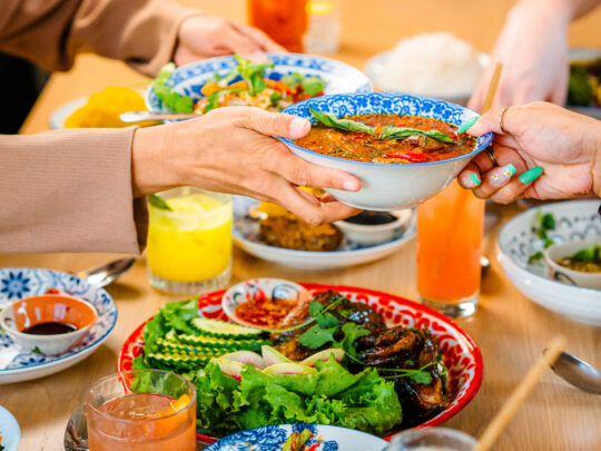 Diners at Kalaya in Philadelphia pass a bowl of vibrant Thai curry across a table filled with colorful dishes, fresh herbs, and bright cocktails.
