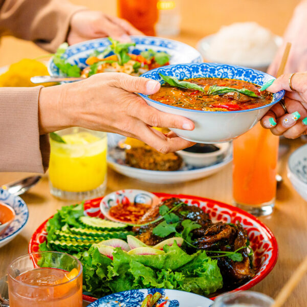 Diners at Kalaya in Philadelphia pass a bowl of vibrant Thai curry across a table filled with colorful dishes, fresh herbs, and bright cocktails.