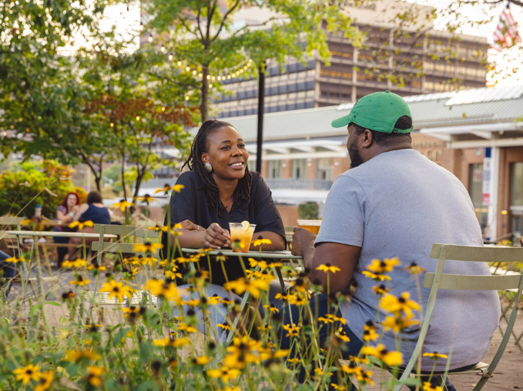 Two people sit at an outdoor table at Liberty Beer Garden in Philadelphia, chatting over drinks surrounded by yellow flowers and string lights in a relaxed garden setting.