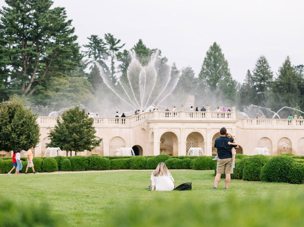 Visitors enjoying the fountains at Longwood Gardens, with a large ornate cream-colored pavilion building, dramatic water jets spraying in the background, manicured hedges, and tall evergreen trees framing the scene.