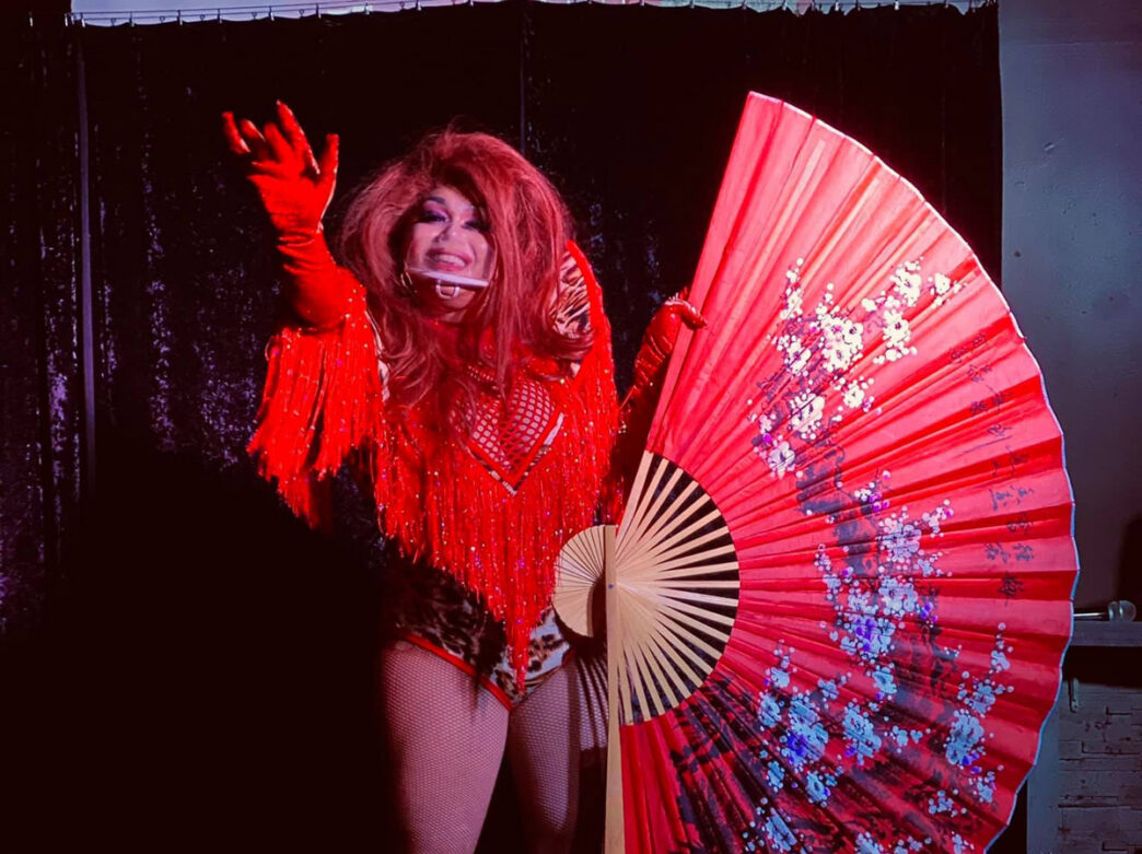 A artista drag Yari Áte faz uma pose confiante no palco usando um vestido de lantejoulas vermelhas com mangas de franjas e um grande leque decorativo com bordados florais como pano de fundo no Lotus Lounge at 254, na Filadélfia.