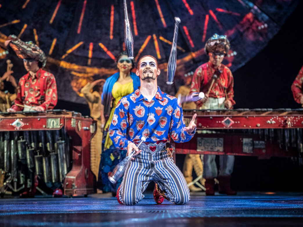 A Cirque du Soleil LUZIA performer juggling silver clubs while kneeling on stage, wearing a colorful blue jacket with red pattern and striped pants, with other costumed performers and musicians visible in the background against an ornate stage set with dramatic lighting.