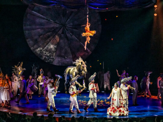 O espetáculo LUZIA do Cirque du Soleil apresenta um elenco diversificado de acrobatas e artistas em trajes coloridos no palco, com um grande adereço de cavalo dourado, um artista aéreo suspenso no alto em um cenário geométrico e uma dramática iluminação de palco em azul e laranja.