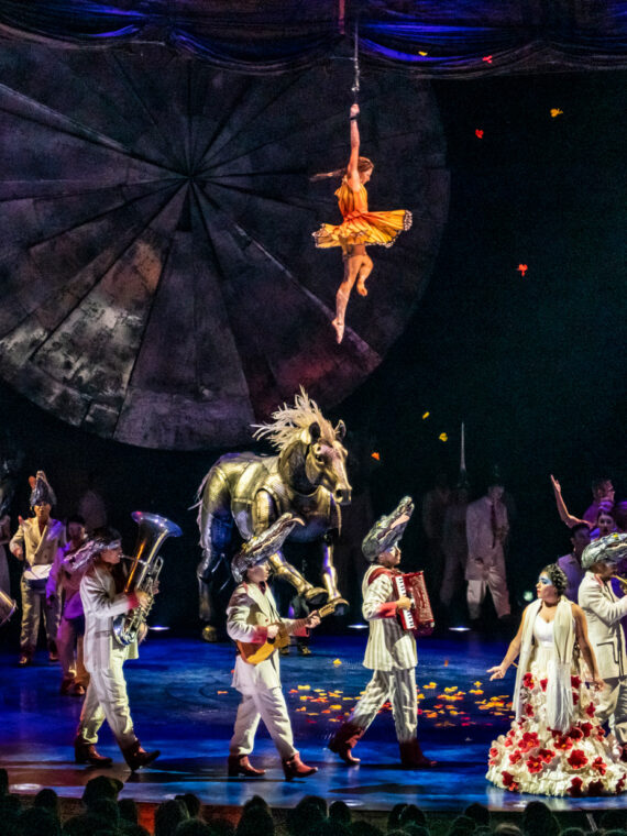 Cirque du Soleil's LUZIA performance featuring a diverse cast of acrobats and performers in colorful costumes on stage, with a large golden horse prop, an aerial performer suspended from above against a geometric backdrop, and dramatic blue and orange stage lighting.