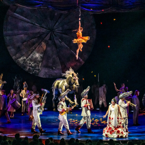 O espetáculo LUZIA do Cirque du Soleil apresenta um elenco diversificado de acrobatas e artistas em trajes coloridos no palco, com um grande adereço de cavalo dourado, um artista aéreo suspenso no alto em um cenário geométrico e uma dramática iluminação de palco em azul e laranja.