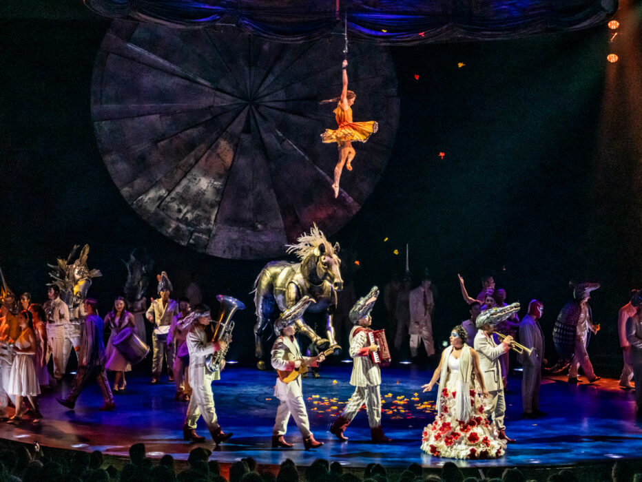 O espetáculo LUZIA do Cirque du Soleil apresenta um elenco diversificado de acrobatas e artistas em trajes coloridos no palco, com um grande adereço de cavalo dourado, um artista aéreo suspenso no alto em um cenário geométrico e uma dramática iluminação de palco em azul e laranja.
