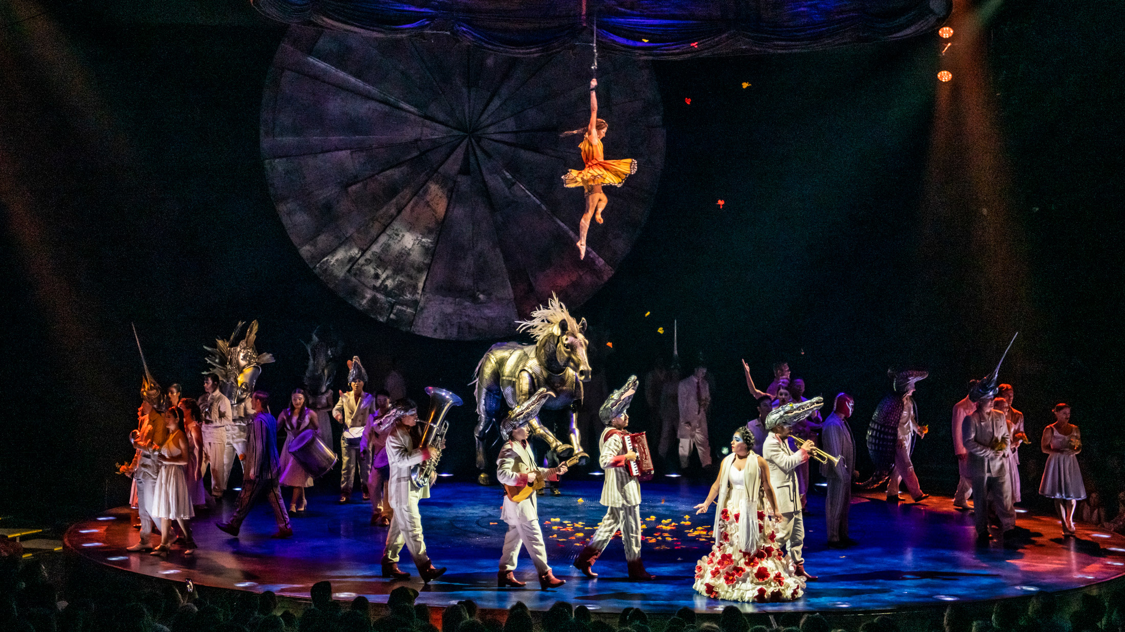 Cirque du Soleil's voorstelling LUZIA met een gevarieerde cast van acrobaten en performers in kleurrijke kostuums op het podium, met een groot gouden paard, een luchtacrobaat die van bovenaf tegen een geometrisch decor hangt en dramatische blauwe en oranje podiumverlichting.