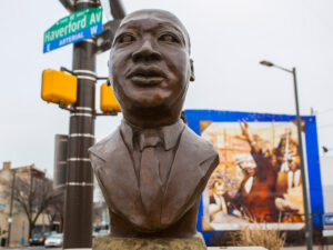 A bronze bust sculpture at the intersection of Haverford Avenue in Philadelphia's Mantua neighborhood, with a large colorful mural visible on a building in the background.