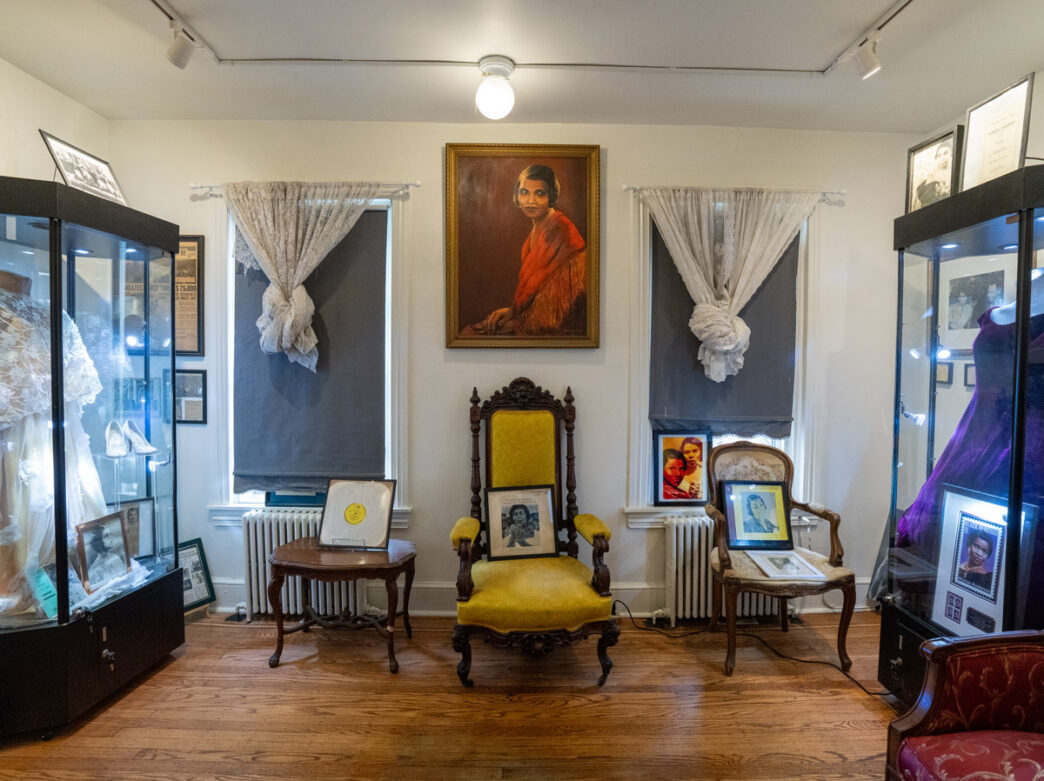 The Marian Anderson Museum displays a large portrait of the celebrated singer above period furniture and antique chairs, with glass cases on either side showcasing her costumes, photographs, and memorabilia from her historic career.