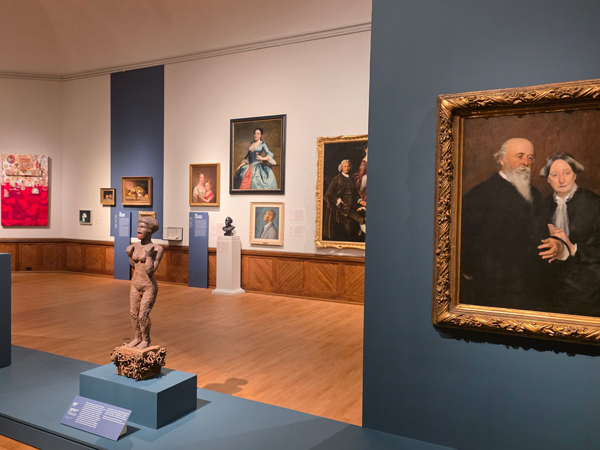 Interior gallery view from Philadelphia’s A Nation of Artists exhibit showing a mix of paintings and sculptures, including a carved wooden figure on a pedestal in the foreground and framed portraits lining the walls.