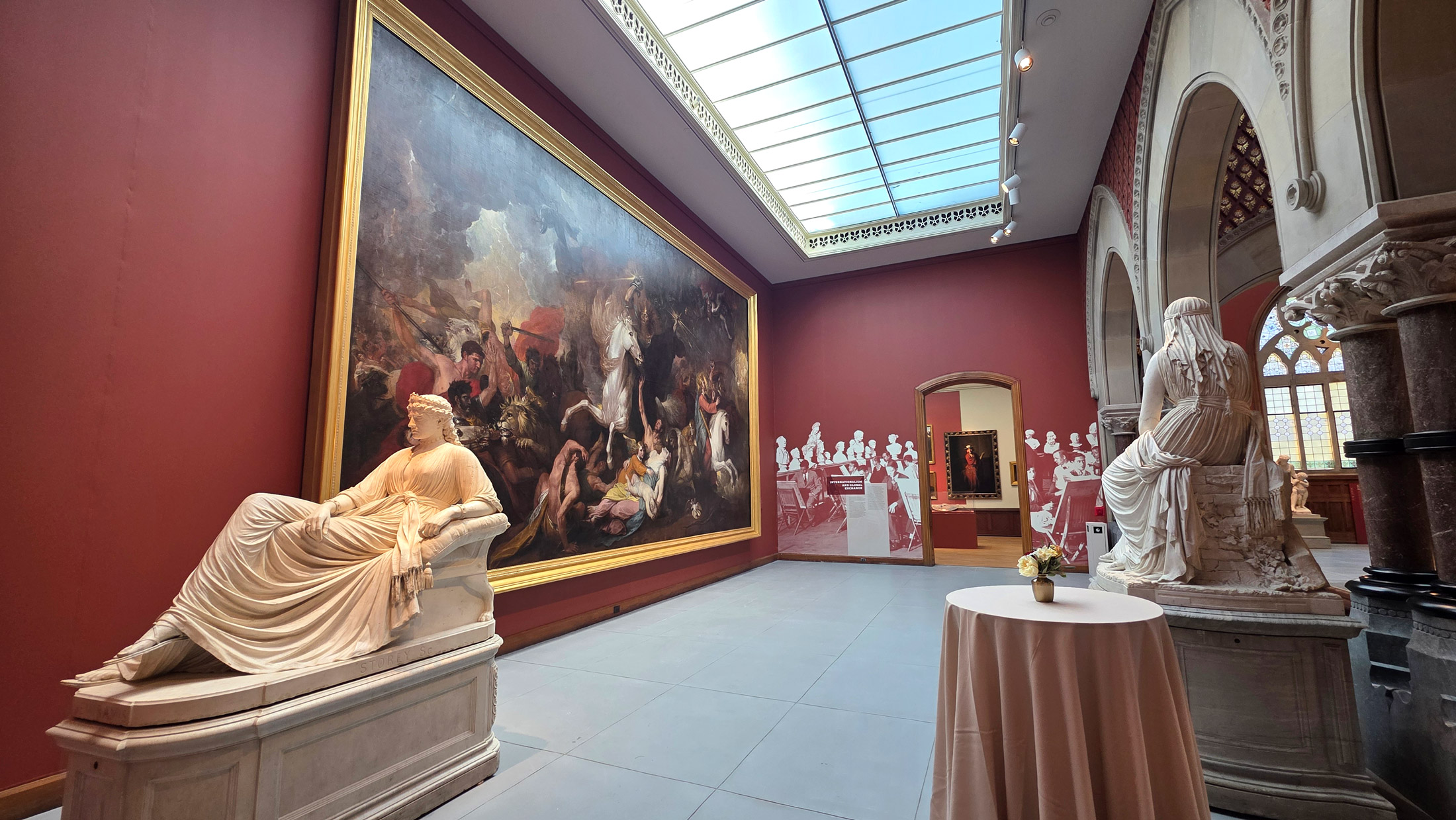 Grand gallery in Philadelphia’s A Nation of Artists exhibit featuring deep red walls, a large dramatic history painting in a gold frame, and classical marble sculptures of seated figures, illuminated by a skylight overhead.