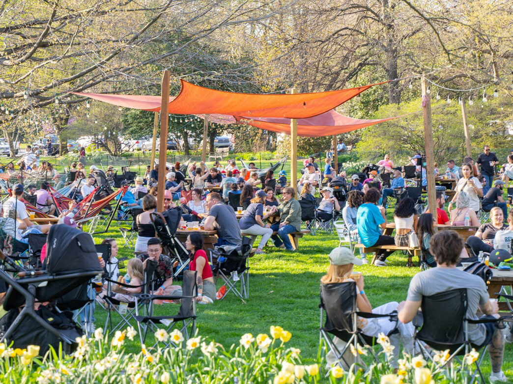 People sit in lawn chairs and at wooden picnic tables under sunshades and string lights at Parks on Tap in Azalea Garden.