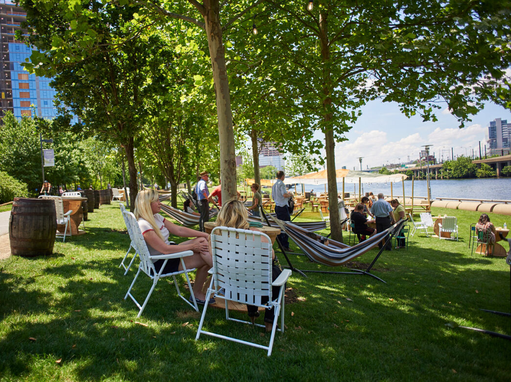 Visitors relax in lawn chairs and hammocks under shade trees at Parks on Tap at Schuylkill River Park in Philadelphia, with the river, tiki-style umbrellas, and the city skyline visible in the background.