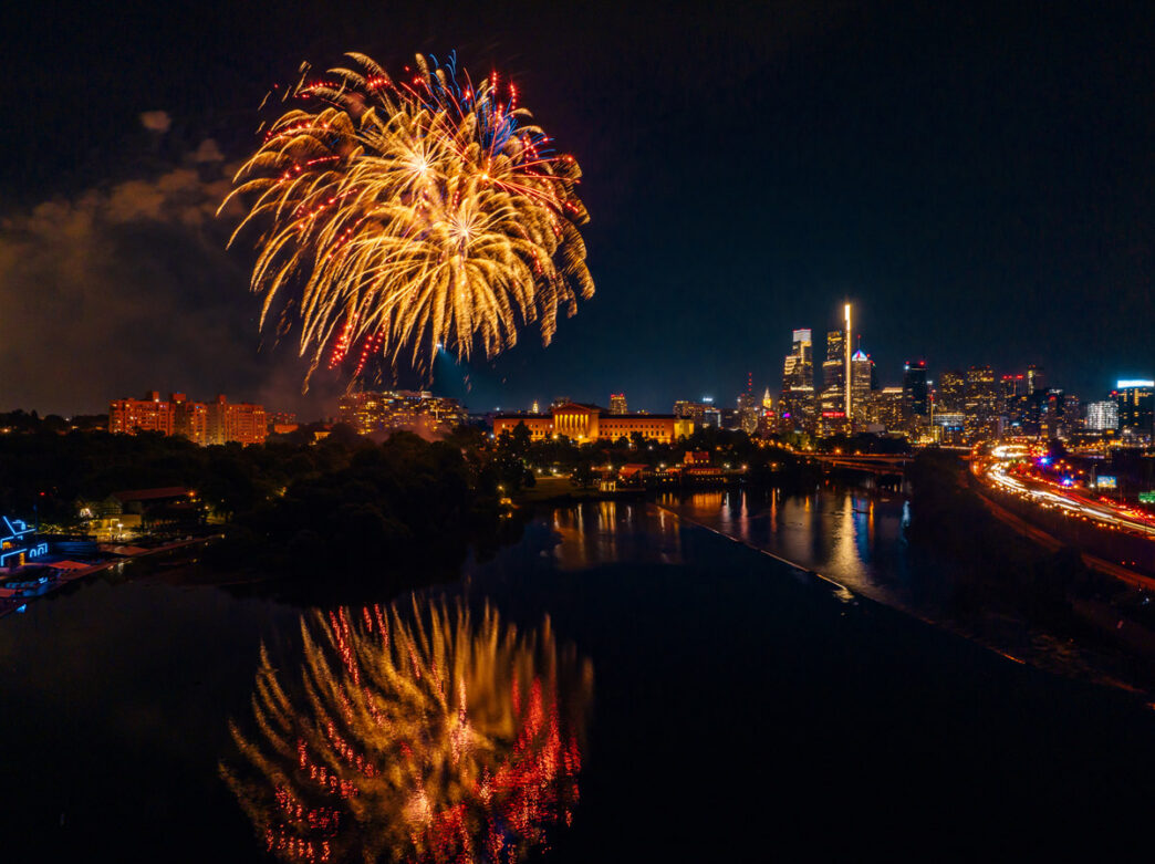 A brilliant fireworks display bursts over the Philadelphia Museum of Art and the Schuylkill River at night, with the city skyline glowing in the background.