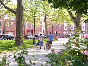Een stel loopt met hun honden langs een zonnig, met bomen omzoomd pad door een park in de wijk Pennsport in Philadelphia. Op de achtergrond zijn bakstenen rijtjeshuizen en auto's te zien die langs de weg geparkeerd staan.