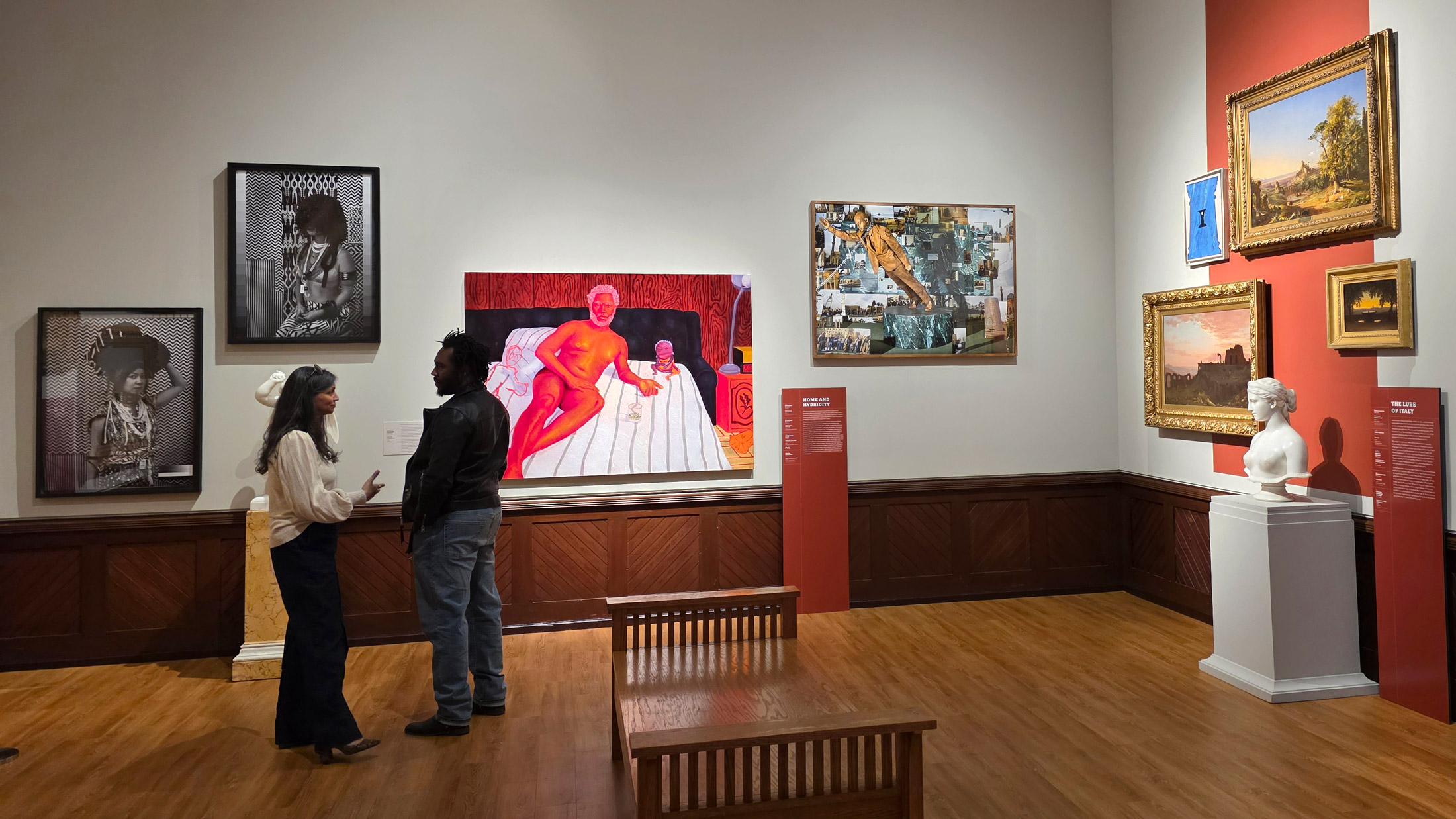 Visitors stand and talk in a gallery of Philadelphia’s A Nation of Artists exhibit, surrounded by a mix of contemporary and classical works, including black-and-white portraits, a vivid red-toned figurative painting, landscape scenes, and a marble bust displayed on a pedestal.