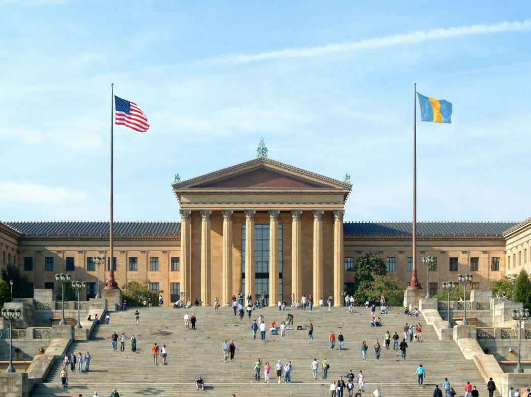 A grande fachada neoclássica do Philadelphia Museum of Art em um dia ensolarado, com visitantes subindo e descendo os icônicos degraus da frente sob a bandeira americana e a bandeira azul e dourada da cidade da Filadélfia.