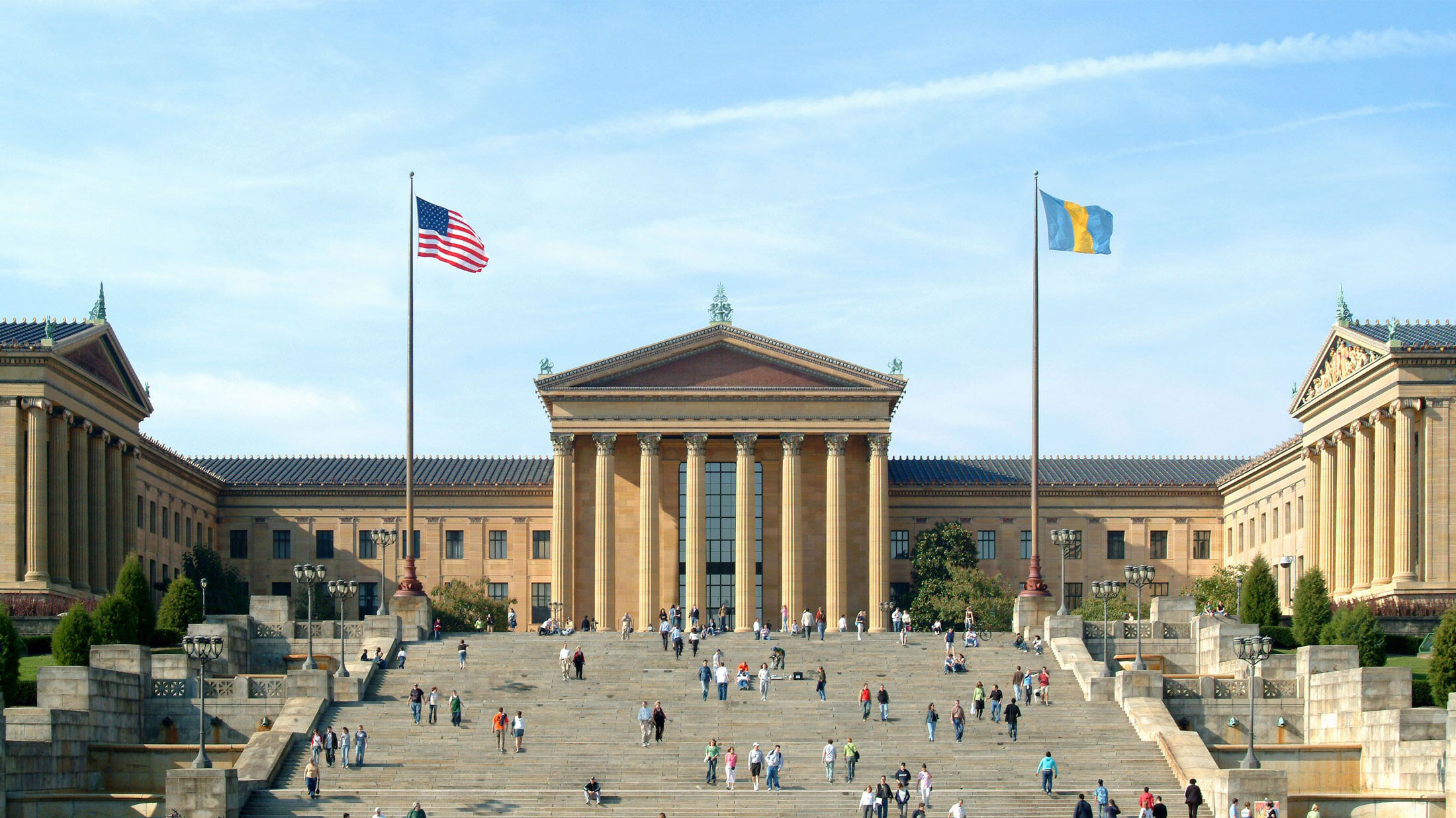 A grande fachada neoclássica do Philadelphia Museum of Art em um dia ensolarado, com visitantes subindo e descendo os icônicos degraus da frente sob a bandeira americana e a bandeira azul e dourada da cidade da Filadélfia.