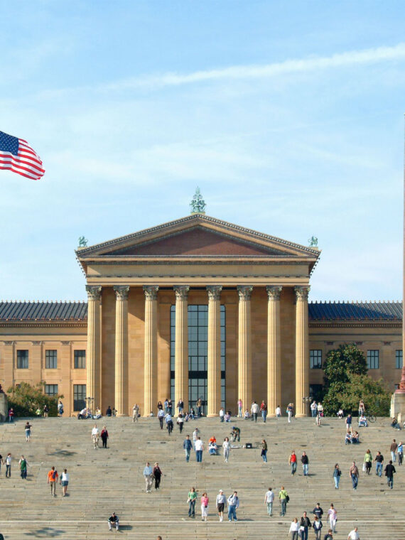 La grande façade néoclassique du Philadelphia Museum of Art par une journée ensoleillée, avec des visiteurs qui montent et descendent les marches emblématiques de la façade sous le drapeau américain et le drapeau bleu et or de la ville de Philadelphie.