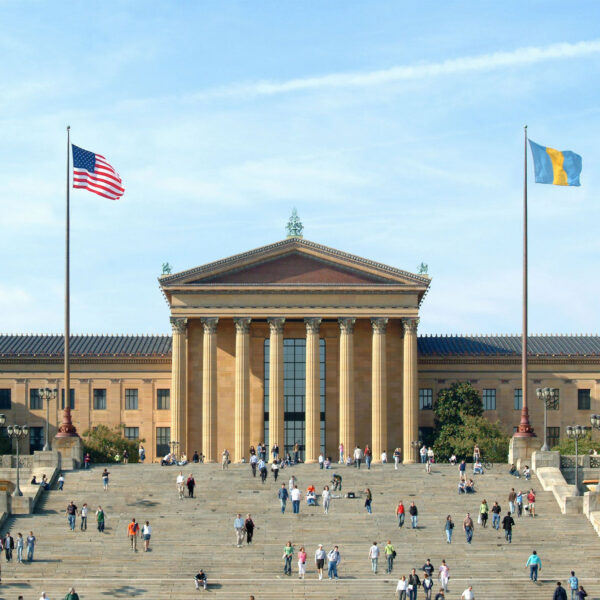 De grootse neoklassieke gevel van de Philadelphia Museum of Art op een zonnige dag, met bezoekers die de iconische trappen op- en aflopen onder de Amerikaanse vlag en de blauwe en gouden Philadelphia stadsvlag.