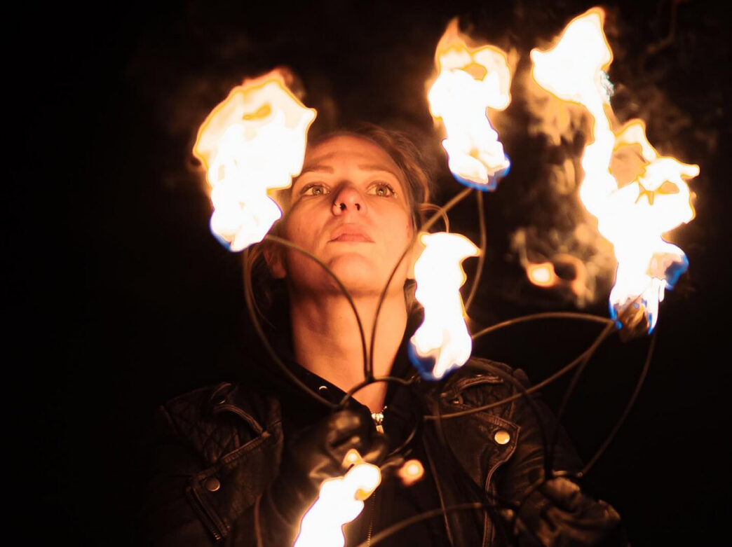 Performer at the Philadelphia School of Circus Arts manipulates multiple flaming torches at night, their face illuminated by the fire against a dark background.