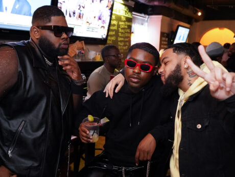 Three people pose together at a lively Philly Black Pride event inside a bar, wearing stylish outfits and sunglasses, with one holding a drink and another flashing a peace sign as people mingle in the background.