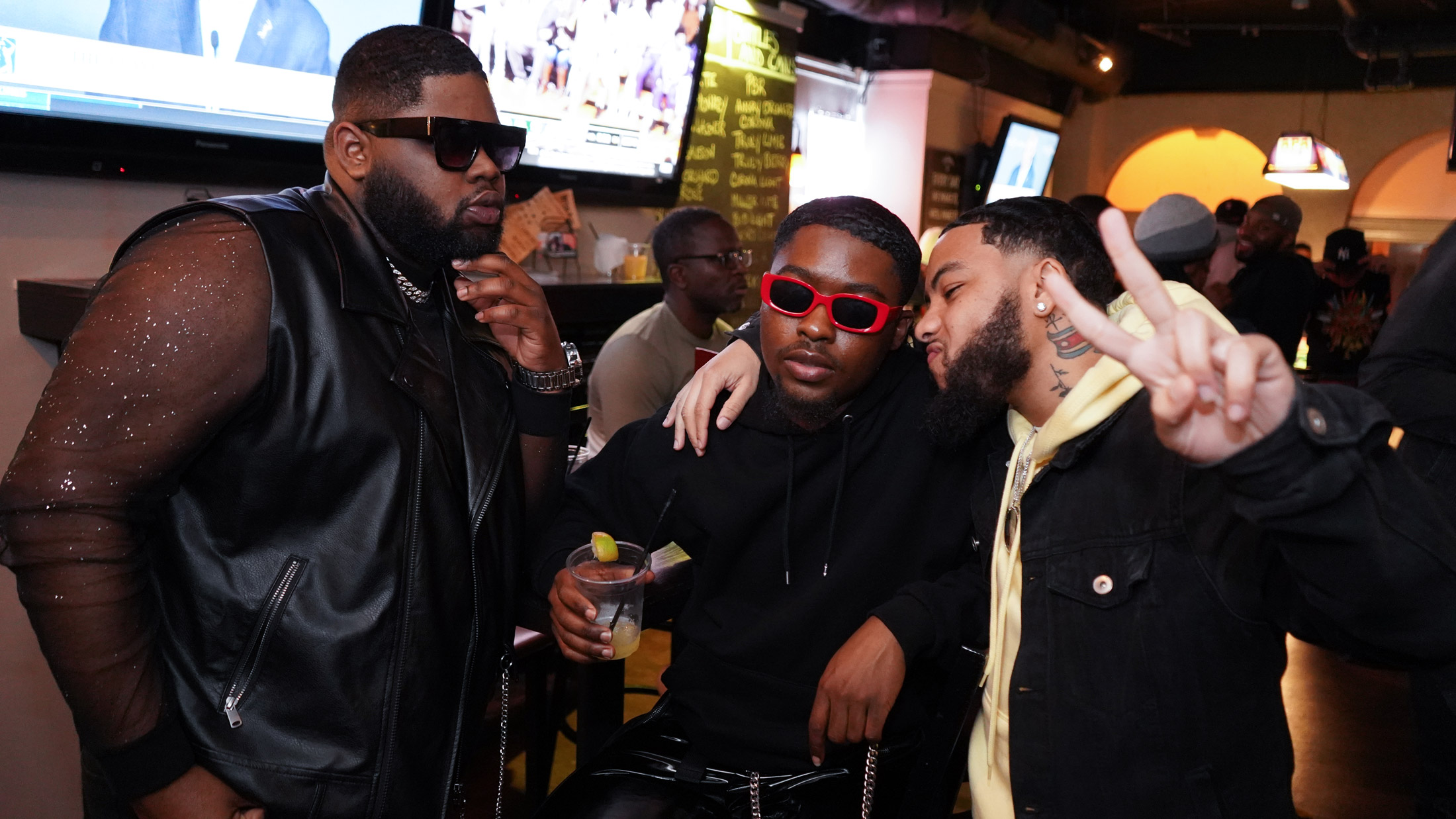 Three people pose together at a lively Philly Black Pride event inside a bar, wearing stylish outfits and sunglasses, with one holding a drink and another flashing a peace sign as people mingle in the background.