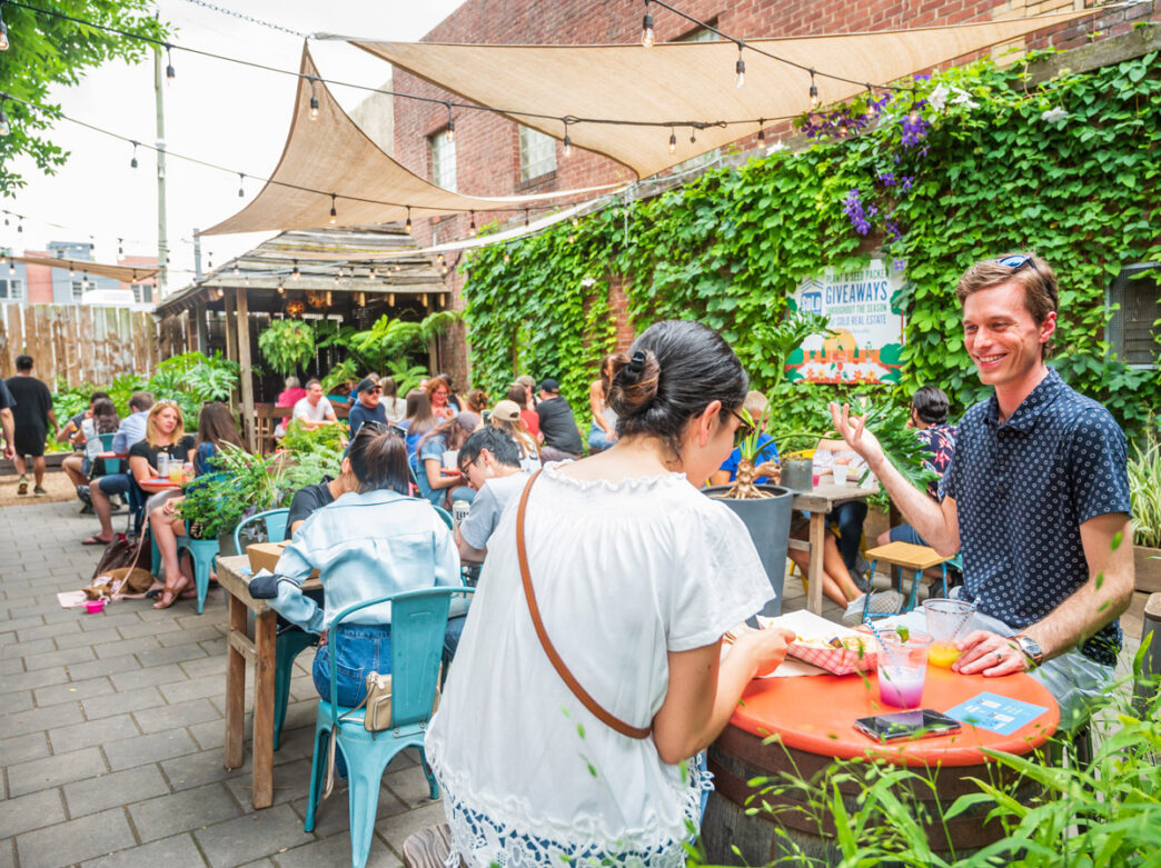 Outdoor scene at the PHS Pop Up Garden on South Street in Philadelphia, with people dining and socializing at colorful tables beneath shade sails, surrounded by lush greenery and string lights along a brick-walled courtyard.