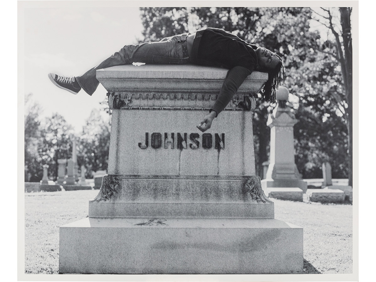 A black and white photograph by artist Rashid Johnson depicting a person lying face-down atop the large granite grave monument of boxer Jack Johnson in a cemetery.