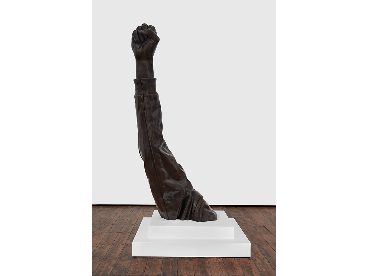A large dark bronze sculpture of a raised arm with a clenched fist, displayed on a white platform in a gallery setting at the Philadelphia Museum of Art.