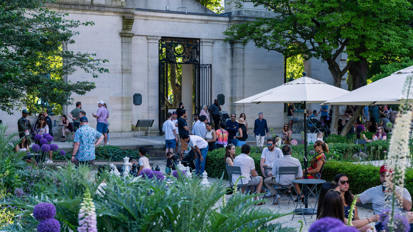 Bezoekers verzamelen zich bij de Garden Bar in het Rodin Museum in Philadelphia, ontspannen aan kleine tafeltjes en onder parasols te midden van weelderig groen, met een stenen tuingang en smeedijzeren hek op de achtergrond.