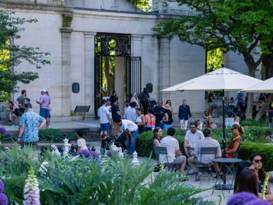 Les visiteurs se rassemblent au Garden Bar du Rodin Museum à Philadelphie, se détendant à de petites tables et sous des parasols au milieu d'une végétation luxuriante, avec une entrée de jardin en pierre et un portail en fer forgé en arrière-plan.