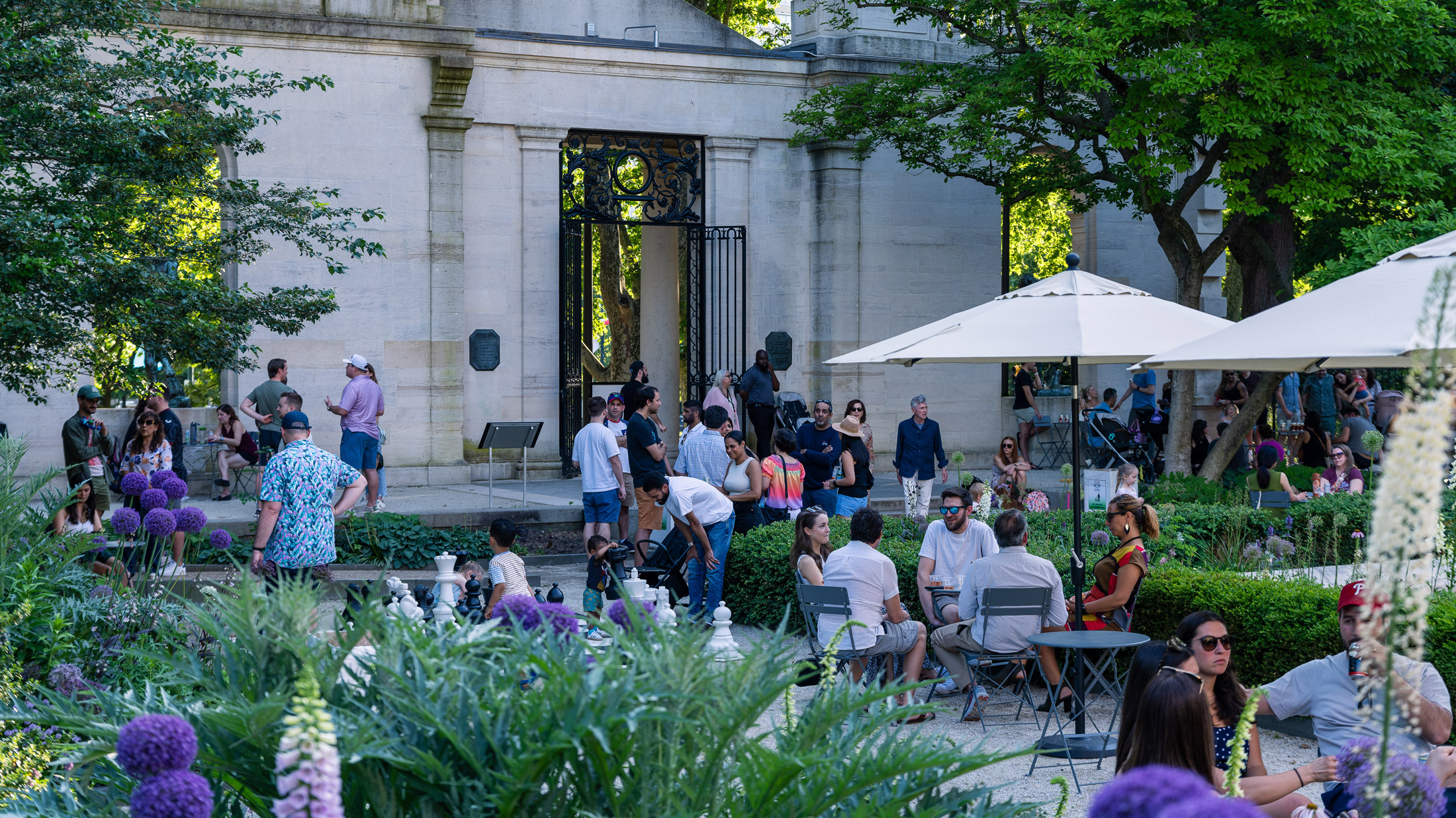Visitors gather at the Garden Bar at the Rodin Museum in Philadelphia, relaxing at small tables and under umbrellas amid lush greenery, with a stone garden entrance and wrought-iron gate in the background.