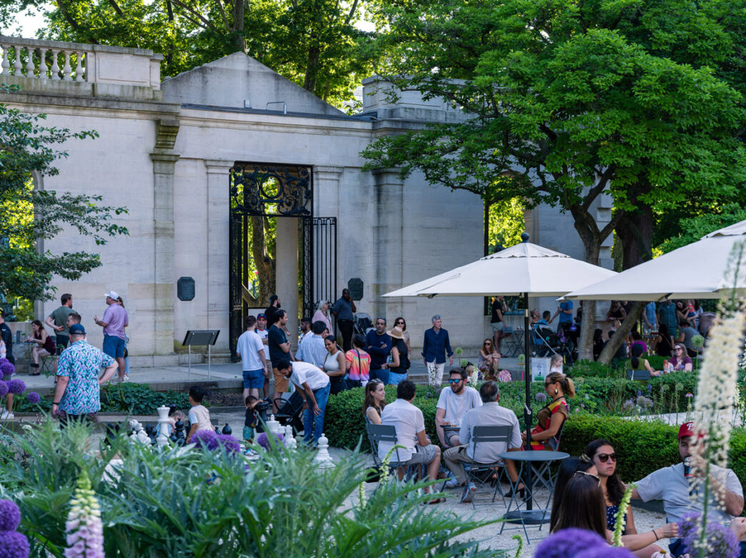 Visitors gather at the Garden Bar at the Rodin Museum in Philadelphia, relaxing at small tables and under umbrellas amid lush greenery, with a stone garden entrance and wrought-iron gate in the background.