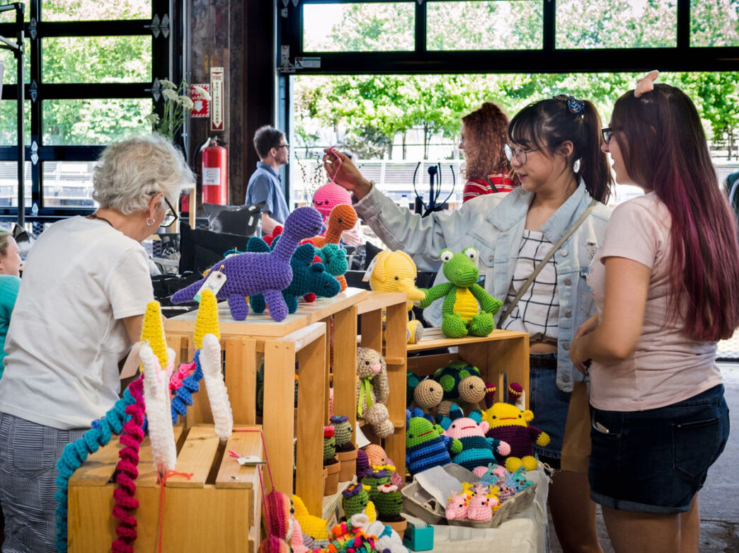 Visitors browsing colorful handmade crocheted items at the Spring Art Bazaar at Cherry Street Pier, including stuffed animals, toys, and decorative pieces in vibrant colors displayed on wooden shelving, with the pier's industrial windows and greenery visible in the background.