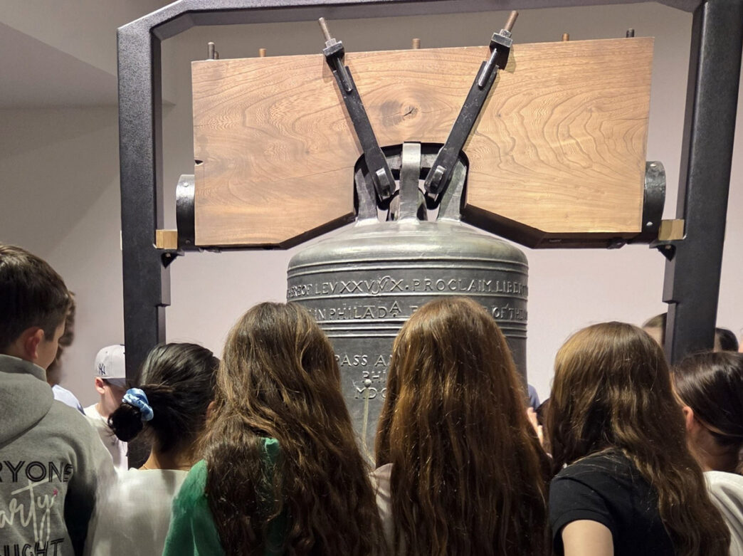 Museum visitors examining a historic bronze bell with wooden mallets in "The Forgotten Freedom" exhibition at the National Liberty Museum in Philadelphia.