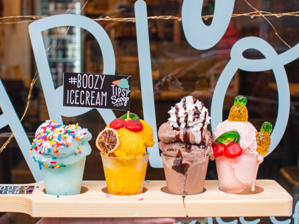 Four decorative boozy ice cream cocktails from Tipsy Scoop with colorful toppings and garnishes displayed in a wooden flight holder.