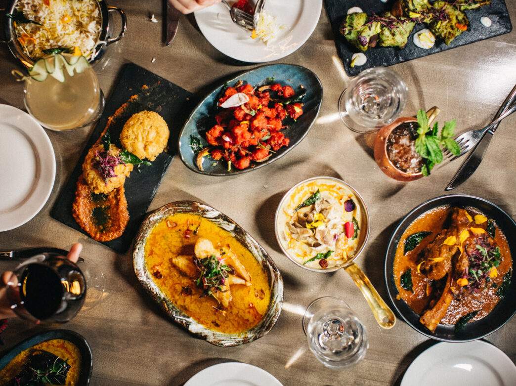 Overhead view of a table at Veda in Philadelphia filled with colorful Indian dishes, including curries, rice, and small plates, alongside cocktails and wine being shared among diners.